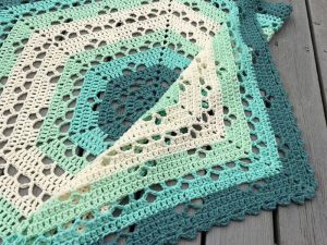 Gros plan d'une couverture au crochet avec un motif géométrique dans des tons de bleu sarcelle, de vert menthe et de crème, exposée sur une surface en bois avec un coin replié - une inspiration parfaite pour les modèles gratuits de couverture au crochet.