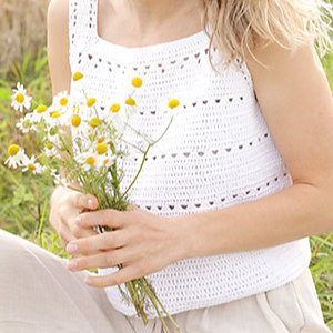 Une personne vêtue d'un débardeur blanc au crochet, inspiré des modèles gratuits de débardeurs au crochet, tient un petit bouquet de marguerites, assise dehors dans un espace vert. Seuls le haut du torse et les mains sont visibles.