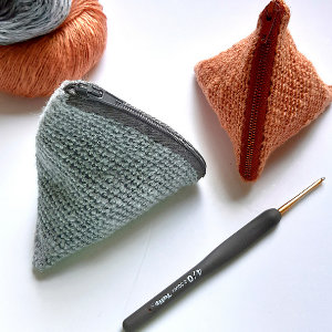 Deux petites pochettes zippées triangulaires texturées, une grise et une orange, sont présentées à côté d'un crochet et de pelotes de fil de couleurs similaires sur une surface blanche, parfaites comme pochette en crochet pour vos essentiels d'artisanat.