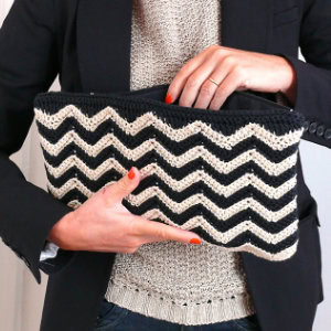 Une personne portant un blazer noir et un pull en tricot beige tient une pochette en crochet à motif zigzag noir et blanc avec les deux mains.