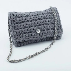 Une petite pochette en crochet grise crochetée à la main avec une bandoulière en chaîne argentée et un fermoir à un seul bouton est présentée sur un fond blanc.