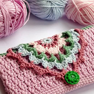 Une charmante pochette en crochet avec un rabat à motif granny-square coloré et un bouton vert, entouré de boules de fil roses et bleues.