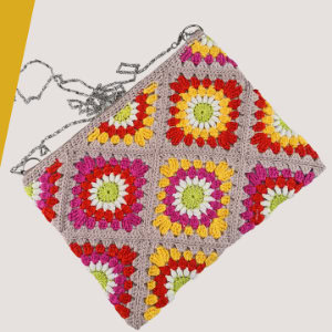 Une pochette en crochet colorée avec des carrés de grand-mère vibrants en rouge, jaune, vert, orange, rose et blanc, avec une bandoulière en chaîne argentée, sur un fond clair avec un accent de coin jaune.