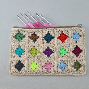 Une pochette en crochet beige avec des motifs carrés colorés et une fermeture éclair, contenant cinq crochets avec des poignées roses dépassant du haut.