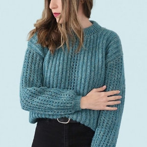 Une femme aux cheveux bruns ondulés portant un pull au crochet texturé bleu sarcelle et un pantalon noir se tient debout sur un fond bleu clair, son bras gauche croisé sur son corps et sa tête tournée sur le côté.