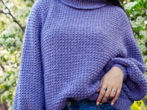 Une personne portant un gros pull au crochet lavande et un jean bleu se tient à l'extérieur, une main dans une poche, sur fond de feuillage vert et d'arbres en fleurs.