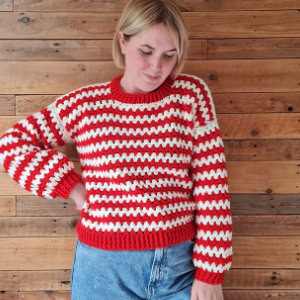 Une personne portant un pull au crochet à rayures rouges et blanches et un jean bleu se tient devant un mur en planches de bois, regardant vers le bas avec une main dans sa poche.