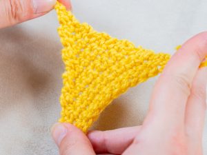 Gros plan de mains tenant un petit triangle jaune tricoté au crochet sur un fond de tissu neutre.