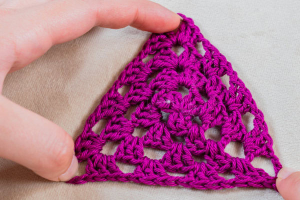 Deux mains tiennent un triangle violet vif au crochet avec un motif ouvert en dentelle, présenté sur une surface en tissu de couleur claire.