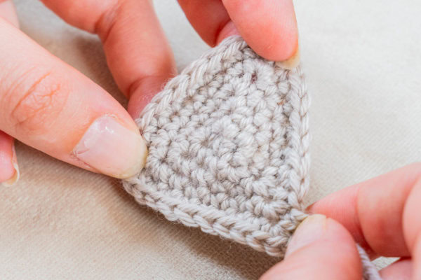 Gros plan de deux mains tenant un petit triangle gris clair au crochet, montrant la texture et les détails du travail du fil sur un fond de tissu neutre.
