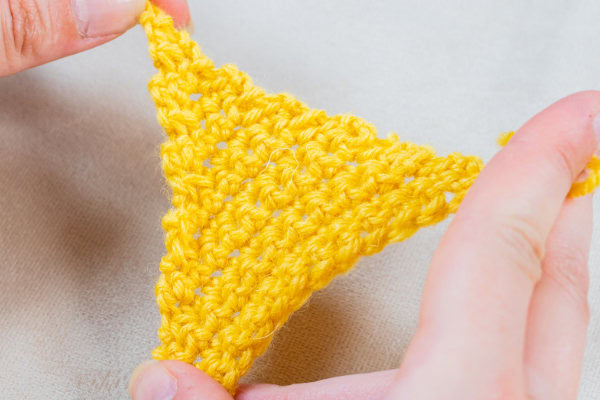 Gros plan de deux mains tenant un petit triangle jaune tricoté au crochet en fil sur un fond de couleur claire.