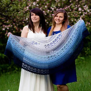Deux femmes se tiennent dehors dans la verdure, souriantes, tenant un grand châle semi-circulaire aux motifs complexes bleu, blanc et bleu marine. Des arbres en fleurs sont visibles en arrière-plan.
