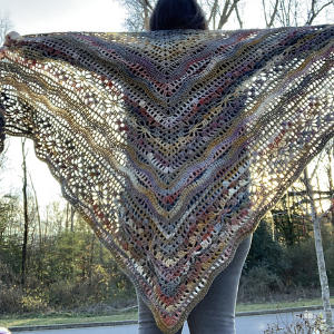 Une personne se tient dehors, tenant un grand châle triangulaire au crochet, orné de motifs multicolores complexes. La lumière du soleil filtre à travers la dentelle ouverte, et des arbres sont visibles en arrière-plan.