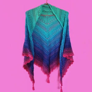 Un châle au crochet dans un dégradé de turquoise, bleu, violet et rose, avec des pompons aux coins, est suspendu sur un fond rose uni.