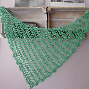 Un châle vert triangulaire au crochet avec un motif ouvert en dentelle est drapé sur une étagère en bois rustique contre un mur violet pâle.