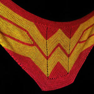 Un châle triangulaire tricoté en rouge et jaune vif, avec un design inspiré de l'emblème emblématique « W » de Wonder Woman sur fond noir.