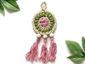 Un ornement en macramé fabriqué à la main inspiré d'un attrape-rêve au crochet présente un centre circulaire vert et rose, une bordure en fil crème, trois pompons roses avec des perles en bois et des accents de feuilles vertes sur fond blanc.