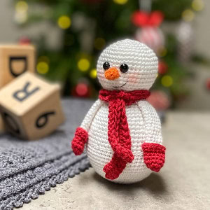 Un bonhomme de neige crocheté, portant une écharpe et des mitaines rouges, se dresse sur une surface grise. Des blocs de bois flous et un sapin de Noël décoré apparaissent en arrière-plan.