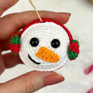 Une main tient un bonhomme de neige crocheté avec des yeux noirs, un nez orange en forme de carotte, un sourire cousu noir et des cache-oreilles rouges et verts. Une boucle dorée permet de le suspendre.