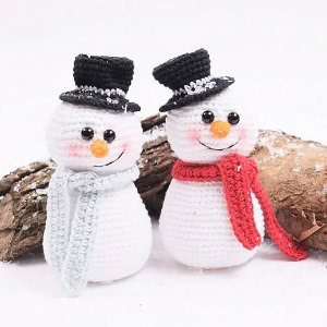 Deux bonhommes de neige crochetés, coiffés de chapeaux haut-de-forme noirs et d'écharpes, l'un bleu clair et l'autre rouge, se tiennent côte à côte sur une surface enneigée, avec une bûche en arrière-plan. Tous deux ont le nez orange, les yeux noirs et les joues roses.