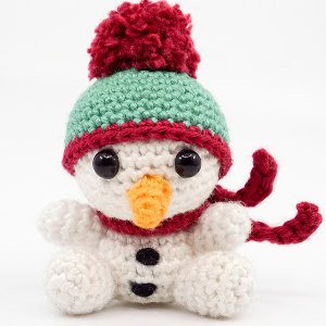 Un bonhomme de neige au crochet avec des yeux noirs en bouton, un nez de carotte orange, des boutons noirs, un chapeau vert avec un pompon rouge et une écharpe rouge, assis sur un fond blanc.