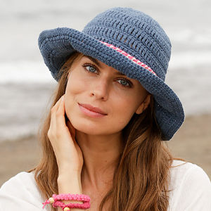 Une femme aux longs cheveux bruns porte un chapeau de soleil bleu crocheté et un haut blanc, souriant doucement. Elle se touche le visage d'une main et porte des bracelets roses au poignet. L'arrière-plan montre une scène de plage floue.