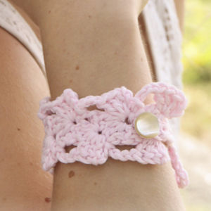 Gros plan sur le poignet d'une personne portant un bracelet rose clair crocheté avec un bouton blanc. Le bracelet présente un délicat motif de dentelle.