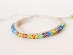Un bracelet fait main au crochet avec un cordon tressé blanc, orné de petites perles de verre colorées dans des tons de bleu, vert, orange et violet, disposées sur une seule rangée.