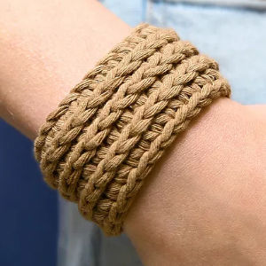 Gros plan d'un poignet orné d'un épais bracelet tressé en corde ou en cordon beige, à l'allure décontractée et naturelle. L'arrière-plan montre une partie d'un vêtement bleu clair.