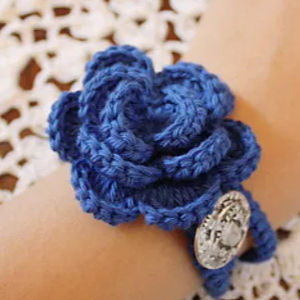 Un bracelet crocheté à fleurs bleues, avec des pétales superposés et une breloque ovale argentée, à porter au poignet. Le fond est un tissu crocheté blanc.