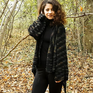 Une femme aux cheveux bouclés, vêtue d'une tenue noire et d'un cardigan en tricot, se tient dans une forêt avec des feuilles mortes, souriant à la caméra.