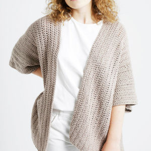 Une personne aux cheveux bouclés porte un cardigan ample marron clair sur une chemise blanche et un pantalon blanc, debout sur un fond clair uni.