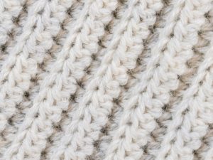 Gros plan d'un tissu tricoté avec un fil épais de couleur crème dans un motif côtelé, ressemblant à des côtes au crochet, montrant une texture douce et des points détaillés.