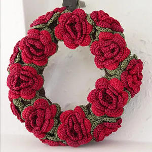 Une couronne circulaire décorée de plusieurs roses rouges crochetées et de feuilles vertes, exposée sur un mur blanc.