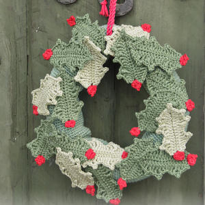 Une couronne faite à la main de feuilles de houx vertes et blanches crochetées, décorées de petites baies rouges, est suspendue à une boucle tressée rouge sur une porte en bois rustique.