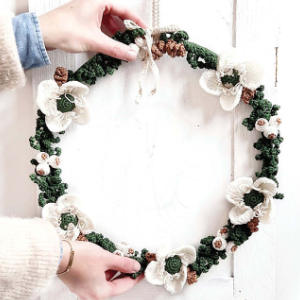 Une personne tient une couronne faite à la main décorée de fleurs blanches au crochet, de feuilles vertes et de petites pommes de pin brunes sur un fond en bois blanc.