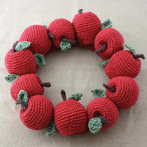 Une couronne composée de pommes rouges crochetées, chacune avec une feuille verte et une tige brune, disposées en cercle sur un fond de tissu neutre.