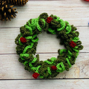 Une couronne crochetée avec des feuilles de fil vert et de petits accents rouges repose sur une surface en bois, avec une pomme de pin visible dans le coin supérieur gauche.