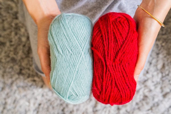 Deux mains tenant deux écheveaux de fil, l'un bleu clair et l'autre rouge, sur une surface grise texturée. La personne, en tenue grise et bracelet doré, est prête à montrer comment faire une couverture au crochet facile.