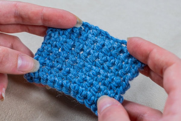 Deux mains tiennent un petit morceau de tissu bleu au crochet, mettant en valeur ses points texturés sur un fond neutre - une inspiration parfaite si vous vous demandez comment faire une couverture au crochet facile.