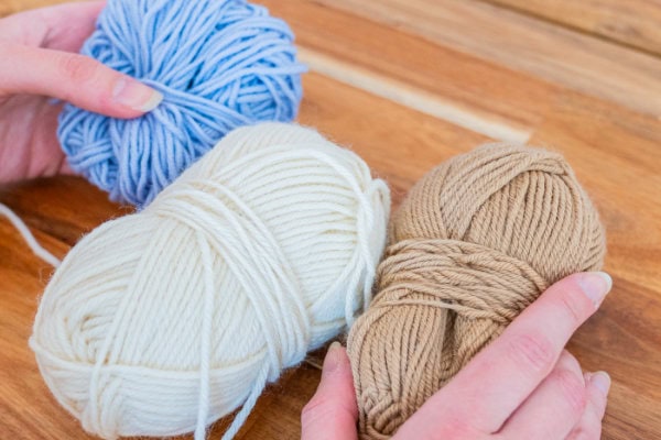Deux mains tiennent trois pelotes de fil bleu, blanc et beige sur une surface en bois - des couleurs parfaites si vous cherchez comment faire une couverture au crochet facile.