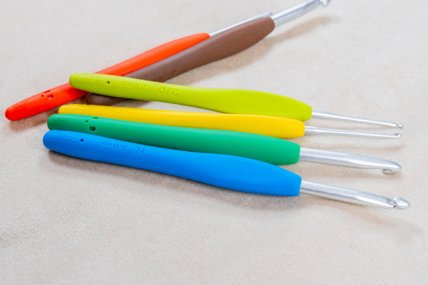 Cinq crochets aux poignées ergonomiques colorées - rouge, vert, jaune, bleu et marron - sont disposés sur une surface beige clair. Les crochets en métal présentent des repères de taille clairs sur chaque poignée ergonomique.