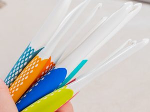 Gros plan d'une main tenant plusieurs crochets ergonomiques colorés avec des poignées texturées et caoutchoutées dans les tons orange, bleu, jaune et sarcelle. Les crochets sont pointés vers le haut sur un fond neutre.