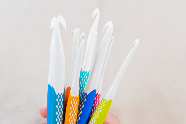 Une main tient plusieurs crochets ergonomiques colorés avec des dessus blancs et des poignées texturées de couleurs vives, présentés sur un fond clair.
