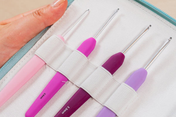 Une main tient un étui contenant quatre crochets ergonomiques colorés avec des pointes en métal et des poignées en caoutchouc dans les tons rose, violet et lilas, chacun étant fixé par un élastique blanc.
