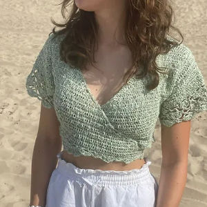 Une femme aux cheveux bruns ondulés se tient sur une plage de sable, vêtue d'un haut court crocheté vert clair et d'un short blanc. Son visage est tourné sur le côté, hors de vue.