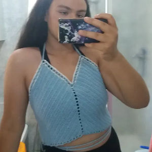 Une personne aux cheveux longs et foncés prend un selfie devant un miroir, vêtue d'un haut dos nu en tricot bleu clair et tenant un téléphone avec une coque sur le thème de l'espace. L'arrière-plan montre une salle de bain.
