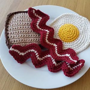Une assiette blanche avec des éléments crochetés ressemblant à une tranche de pain grillé, un œuf au plat et deux tranches de bacon, placés sur une surface en bois.