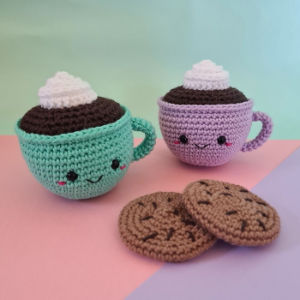 Deux tasses de chocolat chaud crochetées avec des visages souriants et de la crème fouettée sur le dessus, une verte et une violette, sont posées à côté de deux biscuits aux pépites de chocolat crochetés sur un fond pastel.