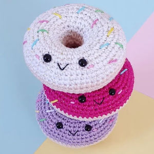 Trois jouets en forme de beignet crochetés avec des visages souriants et des paillettes colorées sont empilés les uns sur les autres sur un fond de couleur pastel.